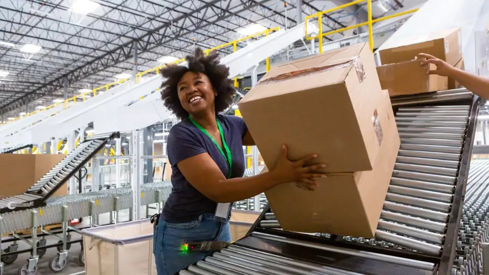 A grinning woman collecting boxes at the bottom of a sorting chute and pushing them along an extendable conveyor belt towards a truck loading dock for delivery