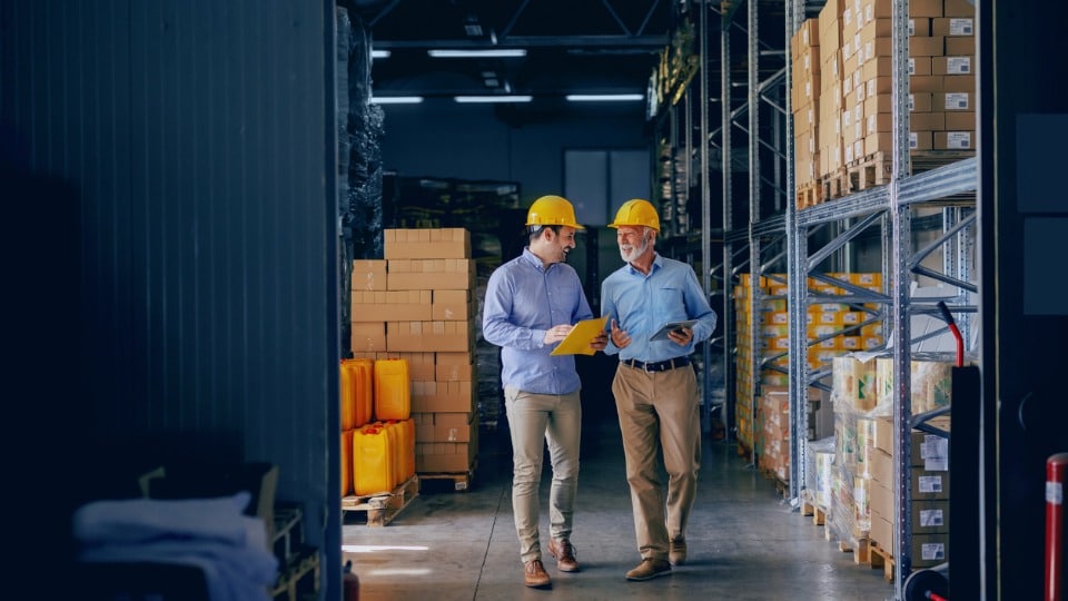 Two business partners in business casual attire and with protective yellow helmets on heads walking and talking in a warehouse