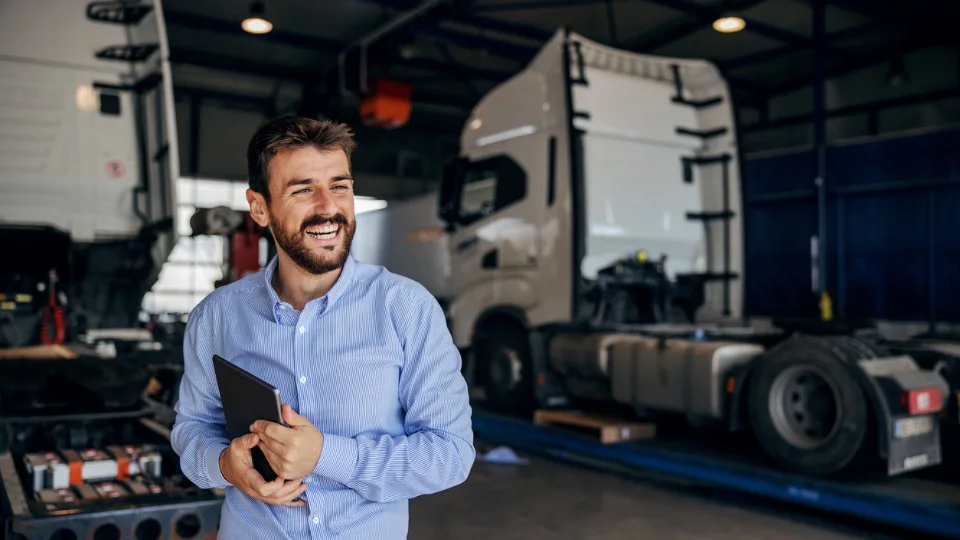 smiling manager standing in a truck garage, holding tablet; in background are trucks.